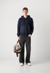 Navy zip-up hoodie with a textured fabric, dark gray oversized jeans, and white sneakers. Beige bag with brown accents held in hand.
