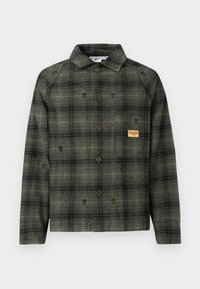 Koszula w oliwkowo-zieloną i czarną kratę z długim rękawem, brązowymi guzikami, kołnierzykiem oraz małą beżową naszywką na kieszeni na piersi.