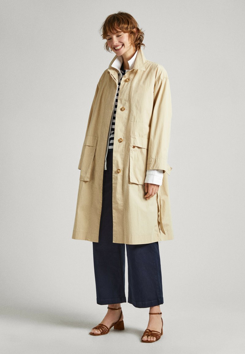 Manteau trench léger beige avec des boutons, deux grandes poches avant et un col montant, associé à un pantalon large noir et des sandales marron.