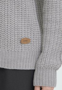 Suéter gris de punto con un patrón texturizado, que cuenta con un dobladillo y puños acanalados. Incluye una pequeña etiqueta de cuero marrón etiquetada "oxmo."