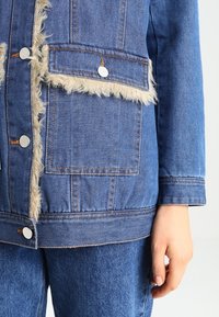 Džínová bunda se syntetickým kožíškem, stříbrnými patentními knoflíky, dvěma předními kapsami a viditelným šitím v středně modré barvě.
