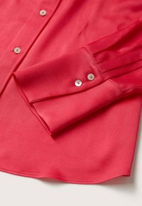 Poignet de chemise rouge à manches longues avec deux boutons blancs et une partie du pied-de-col boutonné sur une surface en tissu lisse.