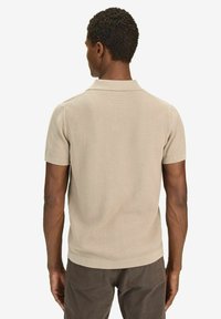 Beige ribbad pikétröja med korta ärmar och platt krage. Har en strukturerad finish och en figurnära silhuett, parat med mörka byxor.