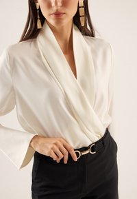 Weiße Satin-Wickelbluse mit weiten Ärmeln, kombiniert mit schwarzen Hosen. Mit einer goldenen, runden Gürtelschnalle als Akzentdetail.