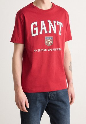 Camiseta roja de manga corta con el texto "GANT", un logo con un escudo de EE. UU. y la frase "American Sportswear", usada por una persona con jeans azules.