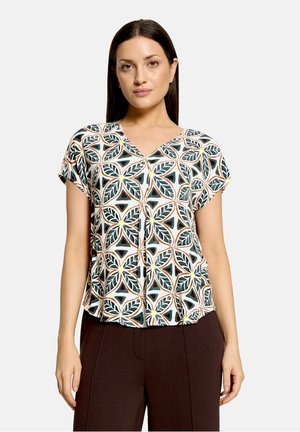 Femme portant un chemisier à manches courtes avec un motif géométrique de feuilles en vert, noir, jaune et marron, associé à un pantalon marron foncé.