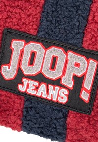 Czerwono-granatowa teksturowana naszywka z napisem "JOOP!" oraz "JEANS" wyszytymi na szaro, otoczona czarnym tłem z czerwonymi akcentami.