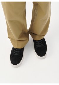 Personne portant un pantalon ample kaki et des baskets noires à semelles blanches, debout sur une surface blanche.