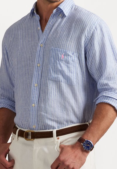 Gai zili svītrota pogu krekla ar krūšu kabatu un rozā logotipu, apvienota ar pelēkām biksēm un brūnu jostu, ar zilu pulksteni.