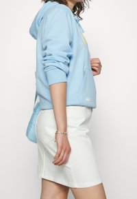 Sweat à capuche bleu clair avec poche kangourou, poignets élastiques et logo ; assorti à une jupe en denim blanche et un sac bandoulière bleu clair.