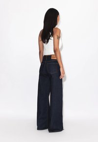 Kvinde med langt mørkt hår iført en hvid ærmeløs top og mørkeblå jeans med vide ben, står vendt mod en hvid væg med en tatovering på armen.