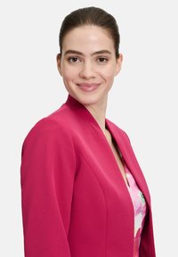 Fuchsia blazer met gestructureerde schouders, gladde textuur en scherpe revers, gelaagd over een bloemenblouse met roze en groene tinten.