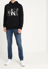 Felpa nera con cappuccio e logo stampato sul davanti, abbinata a jeans blu e sneakers bianche, esposta su un modello in piedi.
