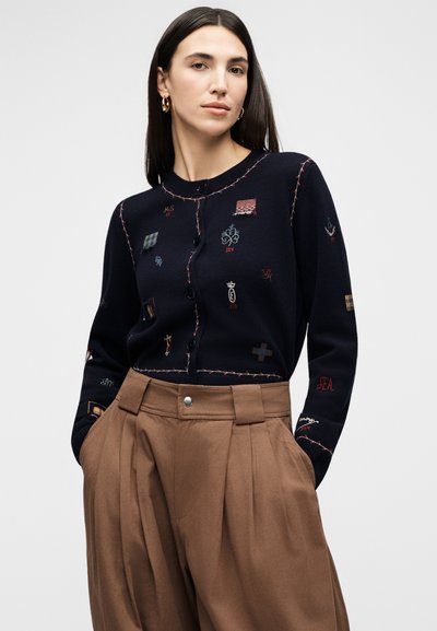 Γυναίκα με μακριά σκουρόχρωμα μαλλιά φοράει μια μπλε ζακέτα με κεντημένες ετικέτες και καφέ ψηλόμεσο πλισέ παντελόνι, στέκεται σε γκρι φόντο.