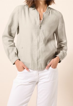 Femme portant une veste zippée beige clair avec des poignets élastiques et un pantalon blanc, les mains posées négligemment dans les poches.