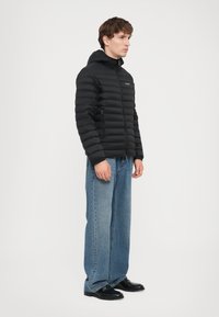 Geacă puf negre cu design capitonat orizontal, glugă atașată, fermoar frontal și buzunare laterale. Asortată cu blugi largi albaștri și pantofi negri.