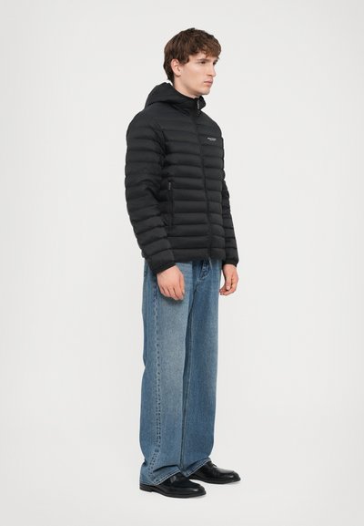 Doudoune noire avec un design matelassé horizontal, capuche attachée, fermeture éclair à l'avant et poches latérales. Associée à un jean bleu ample et des chaussures noires.