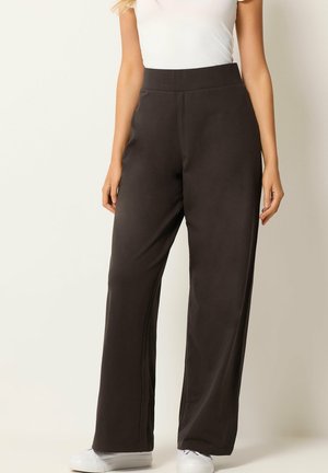 Femme portant un pantalon noir taille haute à jambes larges, un haut blanc à manches courtes et des baskets blanches, debout devant un fond uni.