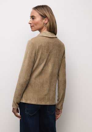 Veste en velours côtelé beige à manches longues et col, présentant une texture côtelée verticale et un ourlet droit, associée à un pantalon en denim foncé.