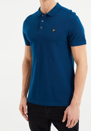 Man wearing a fitted blue short-sleeve polo shirt with buttons and a small yellow bird logo on the chest, paired with black pants.