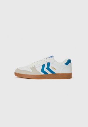 Sneaker blanche avec des accents en suède gris clair et un design en chevron bleu. Dispose d'une semelle en caoutchouc gomme texturée et d'un système de laçage. Marquage doré.