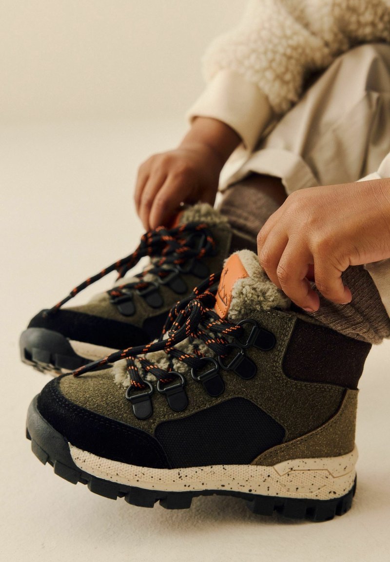 Les mains d’un enfant nouent les lacets noirs et orange de bottes d’hiver vertes et noires avec des semelles beige tachetées et une doublure en polaire.