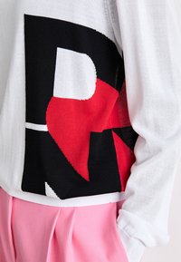 Pull blanc avec un motif géométrique abstrait noir et rouge, associé à un pantalon plissé rose, main dans la poche.