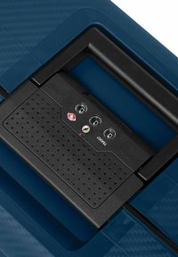 La tapa de la maleta rígida de color azul oscuro presenta un panel de control texturizado en negro con tres diales redondos y una cerradura. Se puede ver un logo rojo.