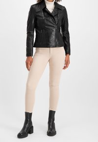 Černá kožená motorkářská bunda s klopovým límcem, zipovými kapsami a přiléhavým střihem, spárovaná s béžovými úzkými kalhotami a černými kotníkovými botami.