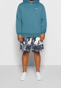 Hombre que lleva puesta una sudadera teal de Calvin Klein, pantalones cortos de rayas blancas y negras, y zapatillas blancas, posando frente a un fondo liso.