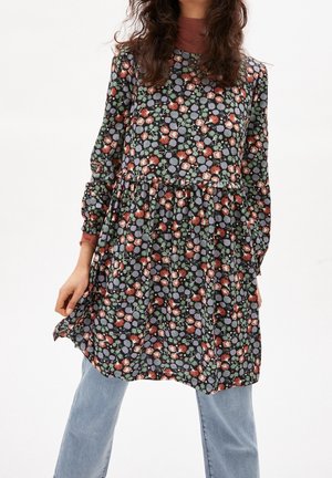 Femme portant une robe noire à manches longues avec des motifs floraux rouges, verts et gris sur un jean bleu clair sur fond uni.