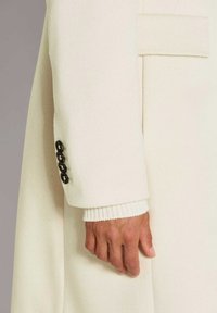 Hand naast lange crèmekleurige mouwen van een jas met vier donkere knopen en een gebreide manchet die eronder zichtbaar is.