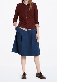 Cardigan lavorato a maglia marrone, camicia a quadri sotto, e una gonna di jeans blu scuro fino al ginocchio. Un paio di scarpe marroni con lacci completano l'outfit.