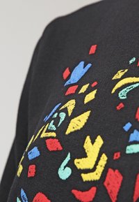Tejido negro con formas geométricas bordadas de colores en azul, rojo, amarillo y verde, con texturas y patrones de puntada variados.