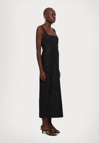 Schwarzes, ärmelloses Midikleid mit strukturiertem Stoff und quadratischem Ausschnitt, kombiniert mit schwarzen, geschnürten Absatzsandalen, präsentiert an einem Model.