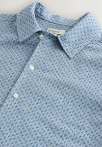 Polo azul claro con un patrón geométrico texturizado, botones blancos y un cuello clásico, colocado plano sobre una superficie blanca.