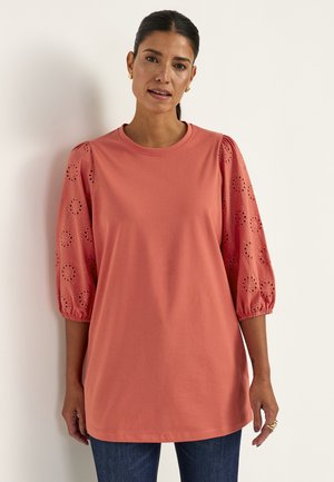 Kvinde iført en koralfarvet tunika med øjlebroderede 3/4 pufærmer og blå jeans, stående foran en ensfarvet lys baggrund.