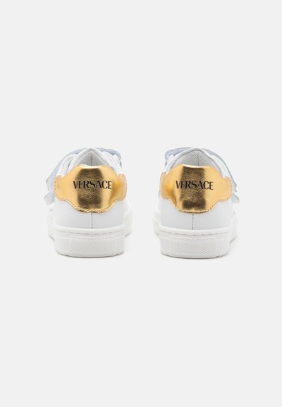 Baskets blanches avec des accents dorés métalliques au niveau du talon et des brides gris clair. Comprend une semelle texturée et le logo "VERSACE" à l'arrière.