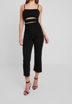 Femme portant un pantalon court noir et un crop top noir sans manches avec une découpe, associé à des sandales noires à talons hauts et à brides.