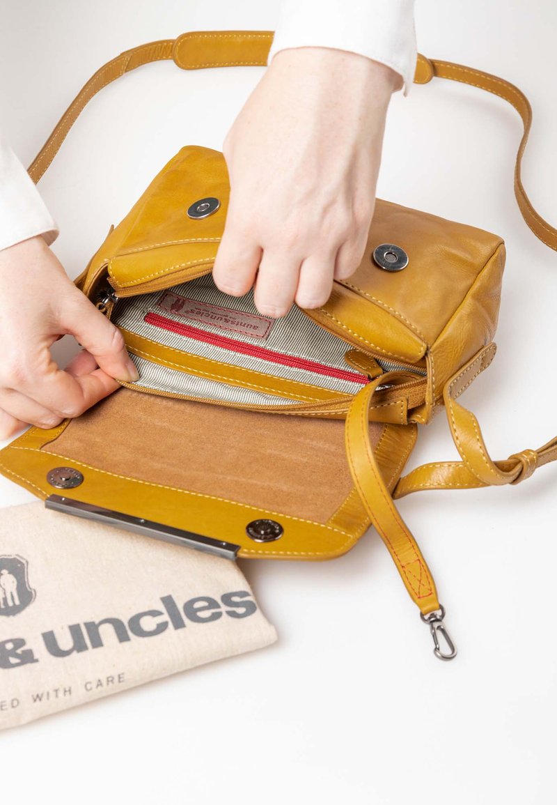 Ledertasche in senfgelb, ausgestattet mit einem Deckel mit zwei Magnetverschlüssen, einem gestreiften Innenfutter und einer Fronttasche mit Reißverschluss.