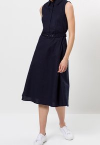 Robe sans manches bleu marine avec un col à revers et une taille cintrée par une ceinture. Confectionnée dans un tissu texturé, elle descend jusqu'au milieu du mollet et présente une silhouette fluide.