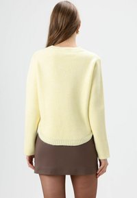 Gele gebreide trui met een afgeronde zoom en geribbelde manchetten, gecombineerd met een bruine rok. Zachte textuur, boxy design, minimalistische stijl.