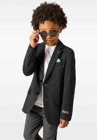 Poisil, kellel on lokkis juuksed, kannab musta bleiserit rohelise tasku ruuduga, halli pükste ja valge särgiga ning reguleerib musti päikeseprille, samal ajal edasi vaadates.