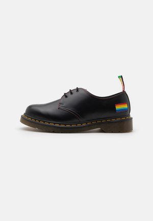Chaussure noire en cuir à lacets avec surpiqûres jaunes, semelle marron, et patch drapeau arc-en-ciel près du talon sur fond blanc.