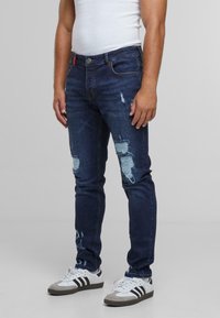 Mørkeblå slitte jeans med lyseblå lapper og rifter, slim fit design, laget av bomull. Kombinert med hvite sko og stripete aksenter.