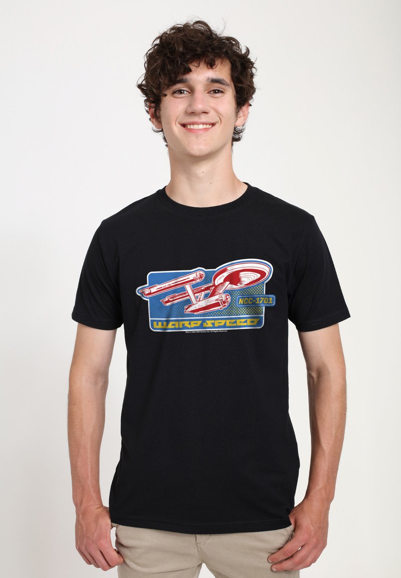 Jeune homme aux cheveux bouclés portant un t-shirt noir avec un vaisseau spatial rouge et l'inscription "NCC-1701 Warp Speed", pantalon beige, souriant.