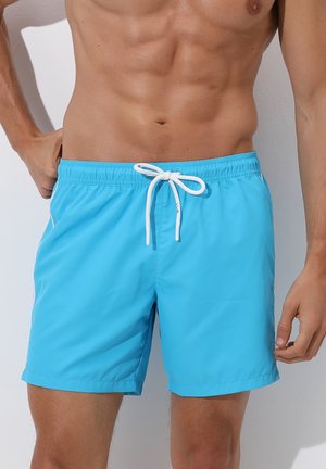Torso maschile che indossa pantaloncini da bagno blu vivace con cordino bianco annodato a fiocco su uno sfondo uniforme.