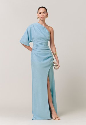 Ženska v svetlo modri obleki z enim ramenom in stranskim razporkom, z zlatimi uhani, zapestnico, ročno torbico in zlatimi sandali z visokimi petami.