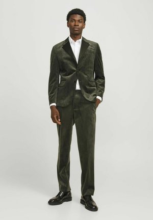 Traje de terciopelo verde con blazer de solapa muestrada, pantalones a juego y una camisa blanca debajo. Unos zapatos de cuero negros completan el conjunto.