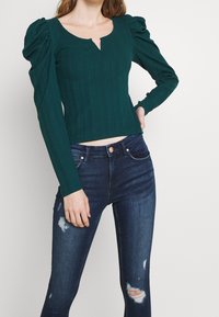 Top de manga larga de canalé en verde azulado con escote en V y hombros abullonados, combinado con jeans oscuros de mezclilla que presentan detalles desgastados y un corte ajustado.